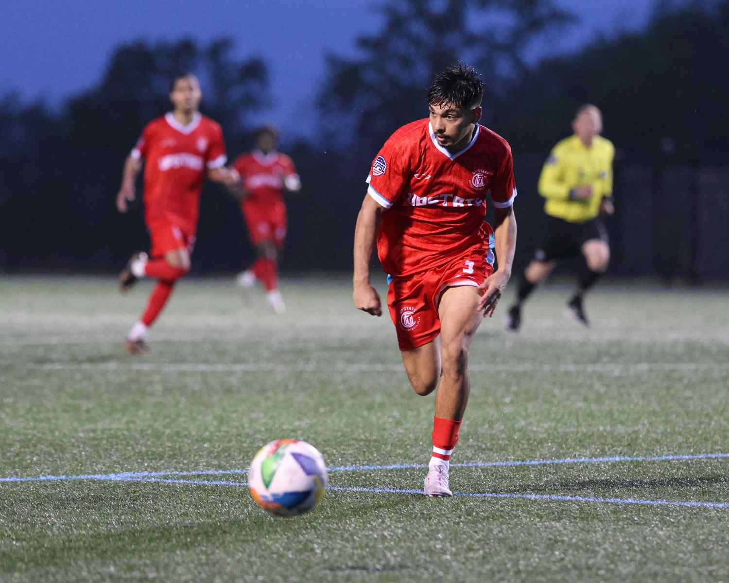 Soccer player in red uniform running on field towards a colorful soccer ball during a nighttime match.