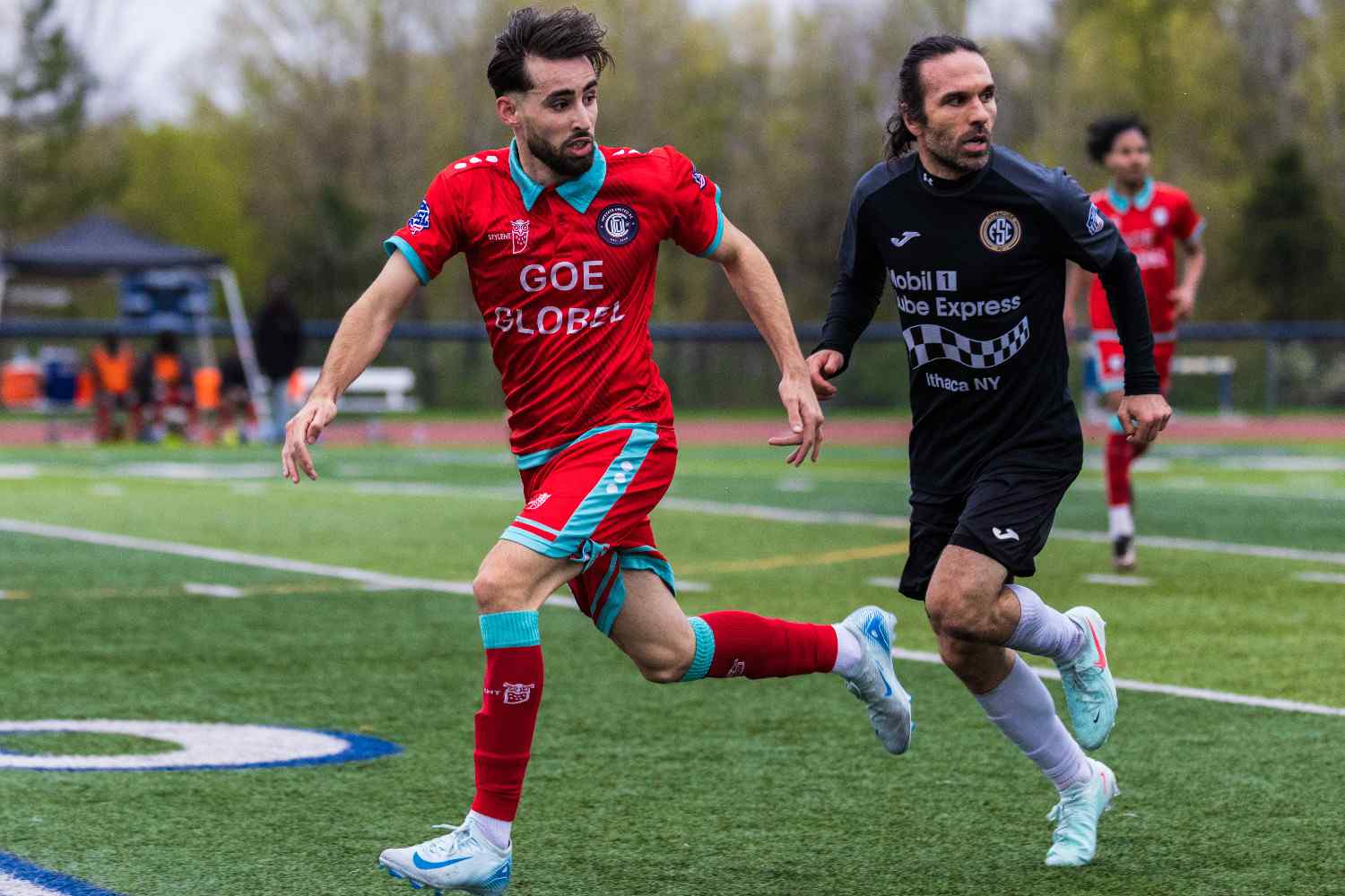 Two soccer players compete on the field, one in a red and turquoise uniform and the other in a black uniform with white socks.