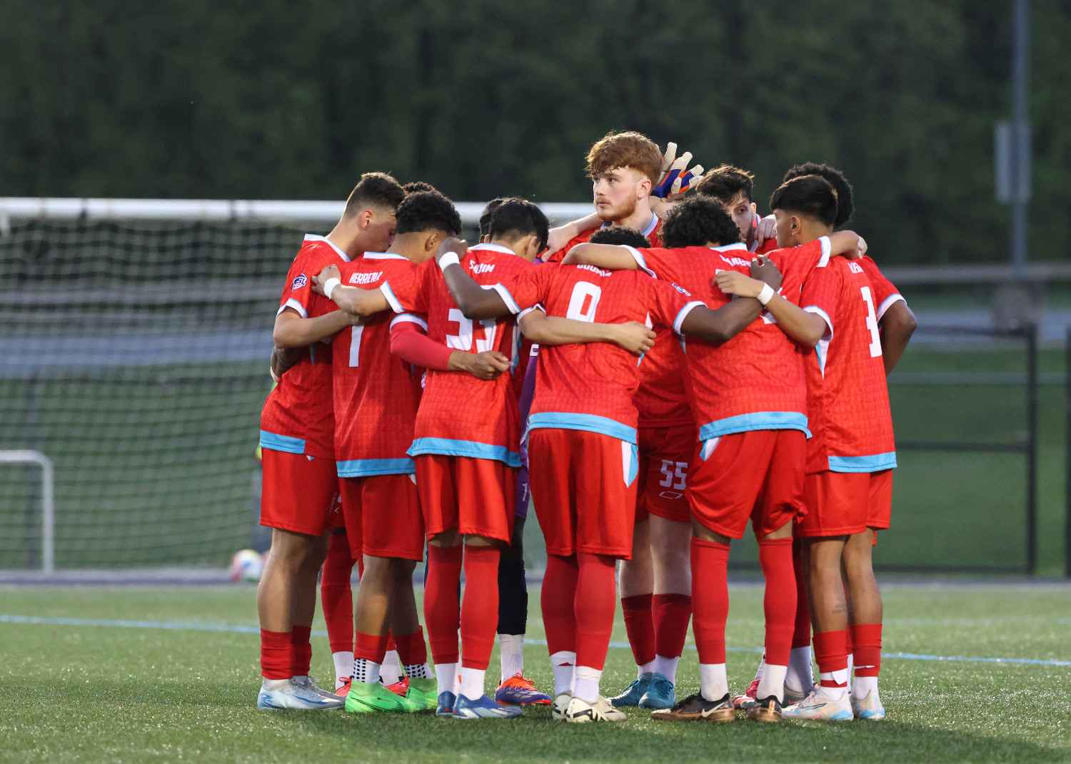 Youth soccer team in red uniforms huddled together on a soccer field near the goal.