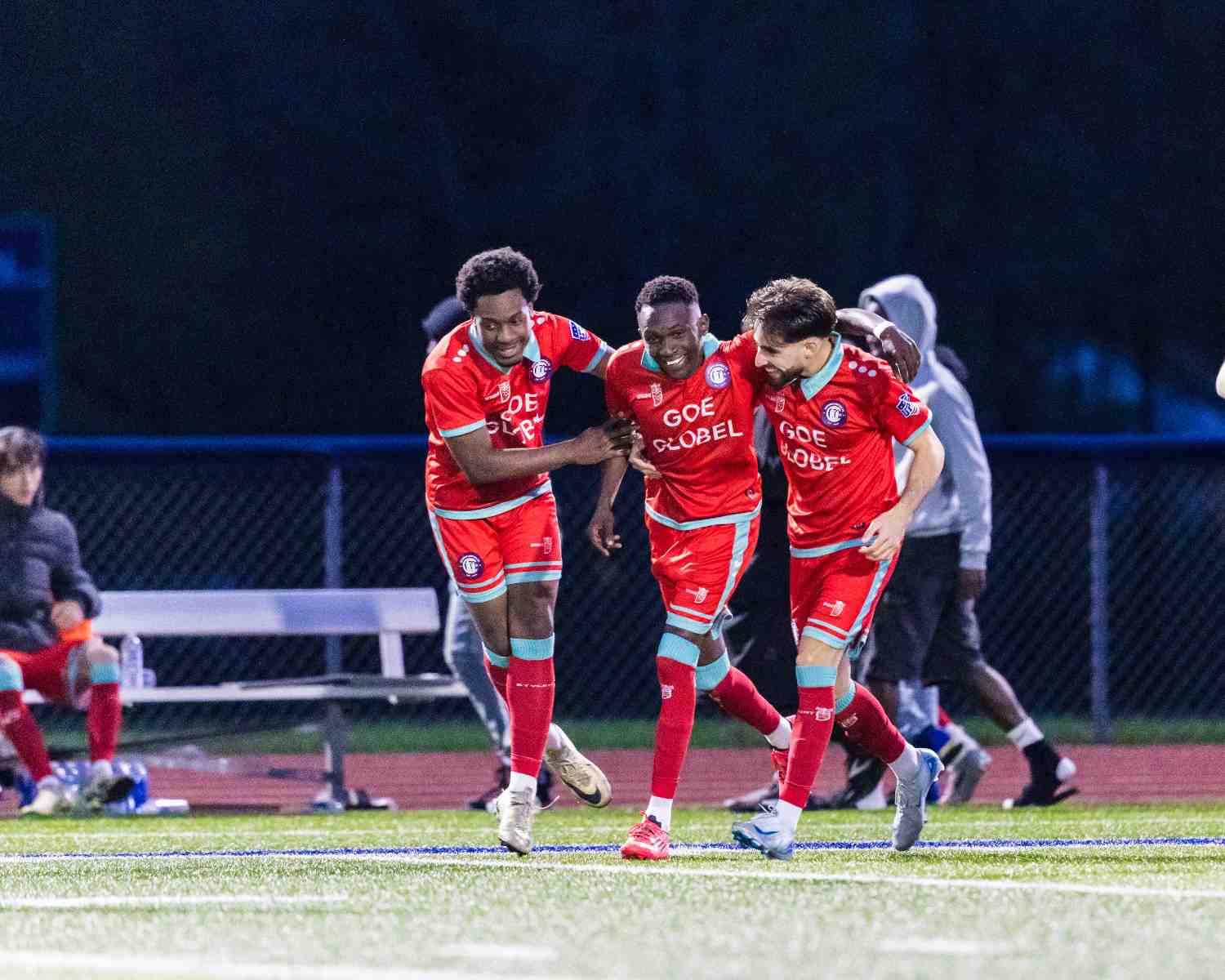 Three male soccer players in red uniforms celebrating on a field at night.