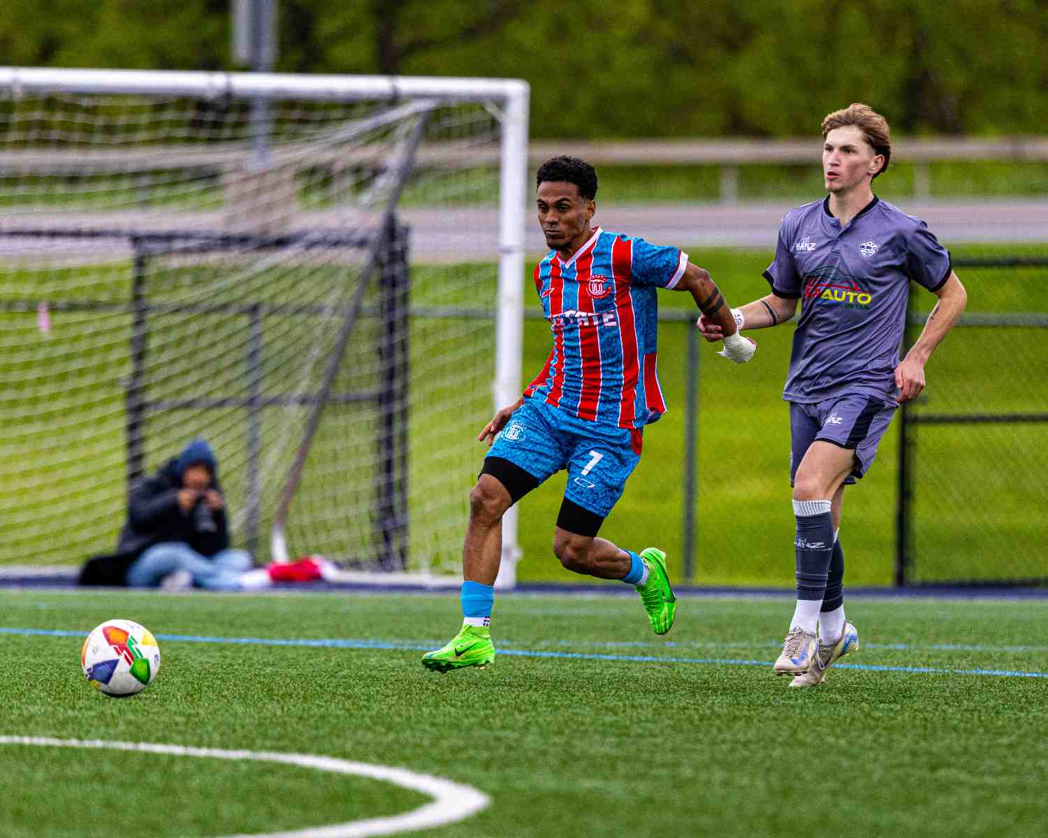 Soccer player in blue and red striped uniform running towards the ball with another player in a purple uniform chasing him on green field.