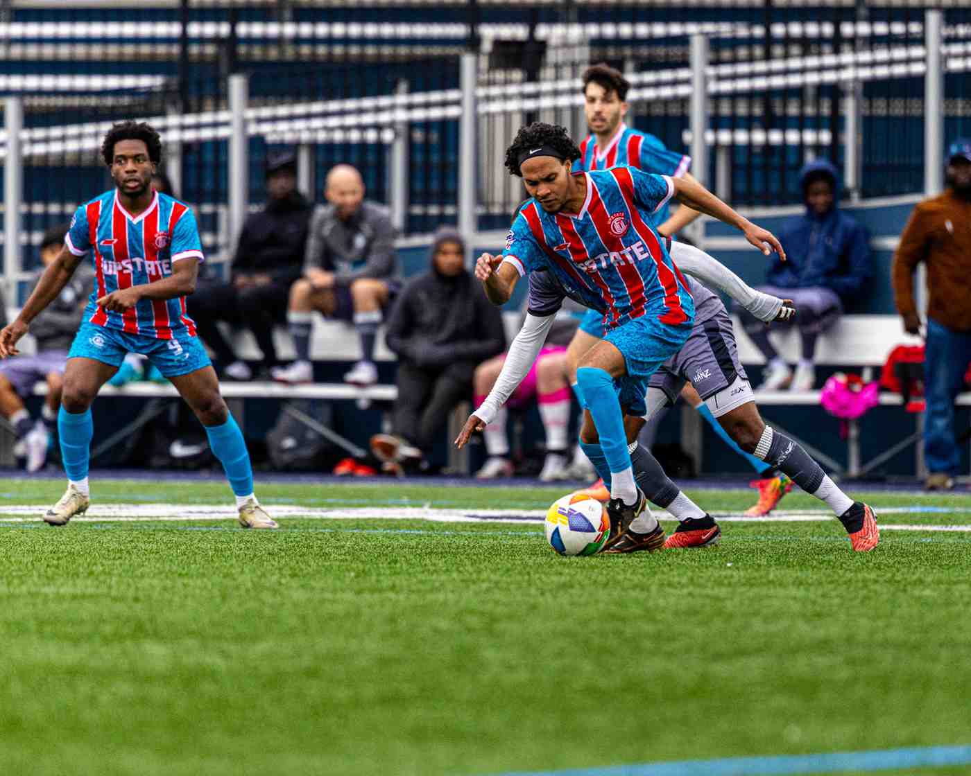 Soccer players in blue and red striped jerseys competing for the ball on a green field with spectators in the background.