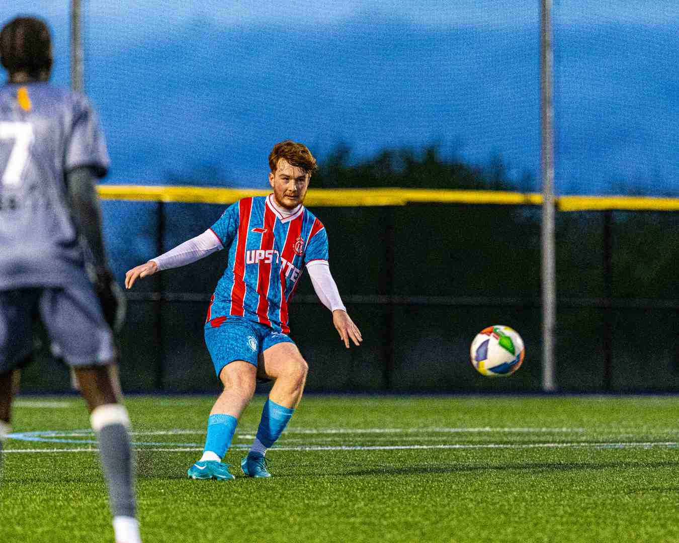 Soccer player in blue and red striped jersey kicking a multicolored ball on a turf field during evening.