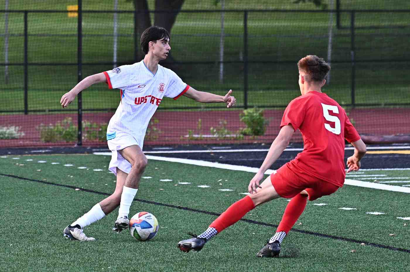 Two male soccer players competing for the ball on a turf field, one in a white Upstate jersey and the other in a red jersey with number 5.