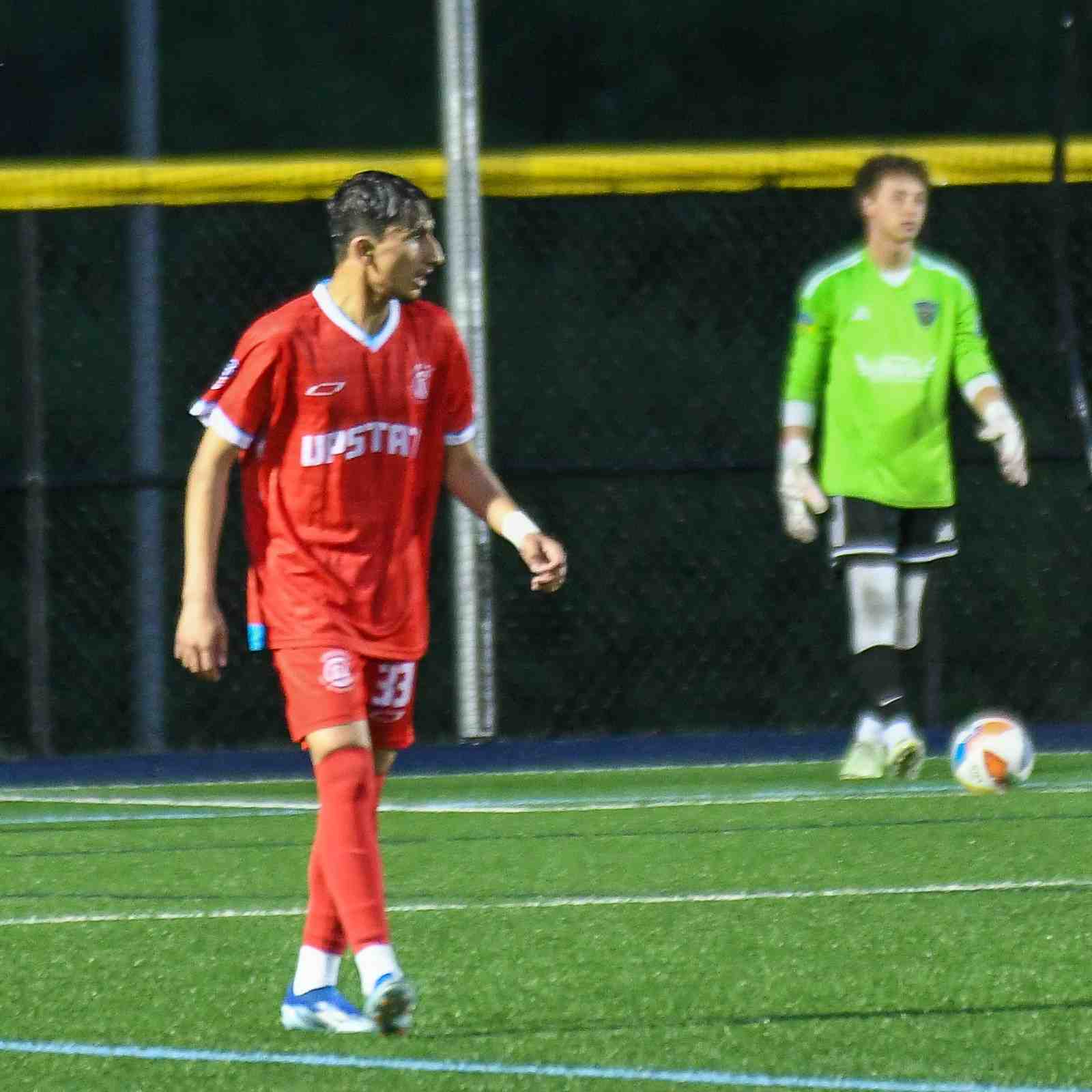 Soccer player in red uniform walking on field with goalkeeper in green jersey and white gloves standing near soccer ball.