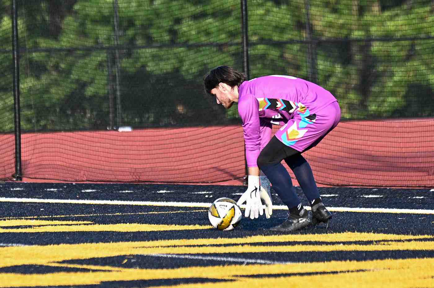 Soccer goalkeeper in a colorful purple uniform bending down to catch a soccer ball on a field with yellow and blue markings.