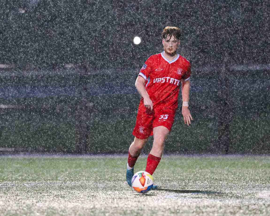 Soccer player wearing a red Upstate uniform playing in heavy rain on a field.
