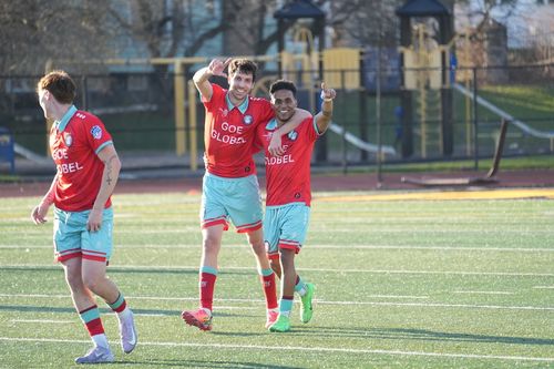 Two soccer players in red and light blue uniforms celebrating on a green field, one with arm around the other pointing forward.