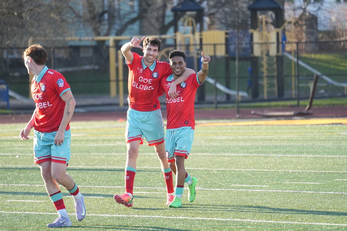 Two soccer players in red and light blue uniforms celebrating on a green field, one with arm around the other pointing forward.