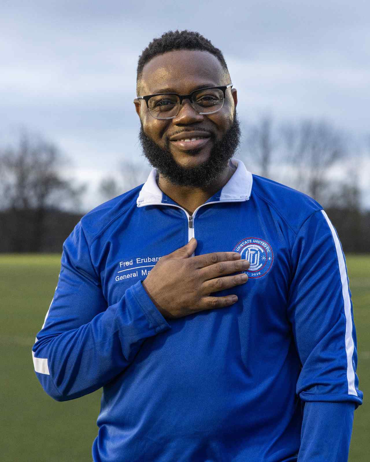 Smiling man with glasses and beard wearing a blue sports jacket with a logo and the name Fred Erubari with title General Manager, hand placed on his chest.