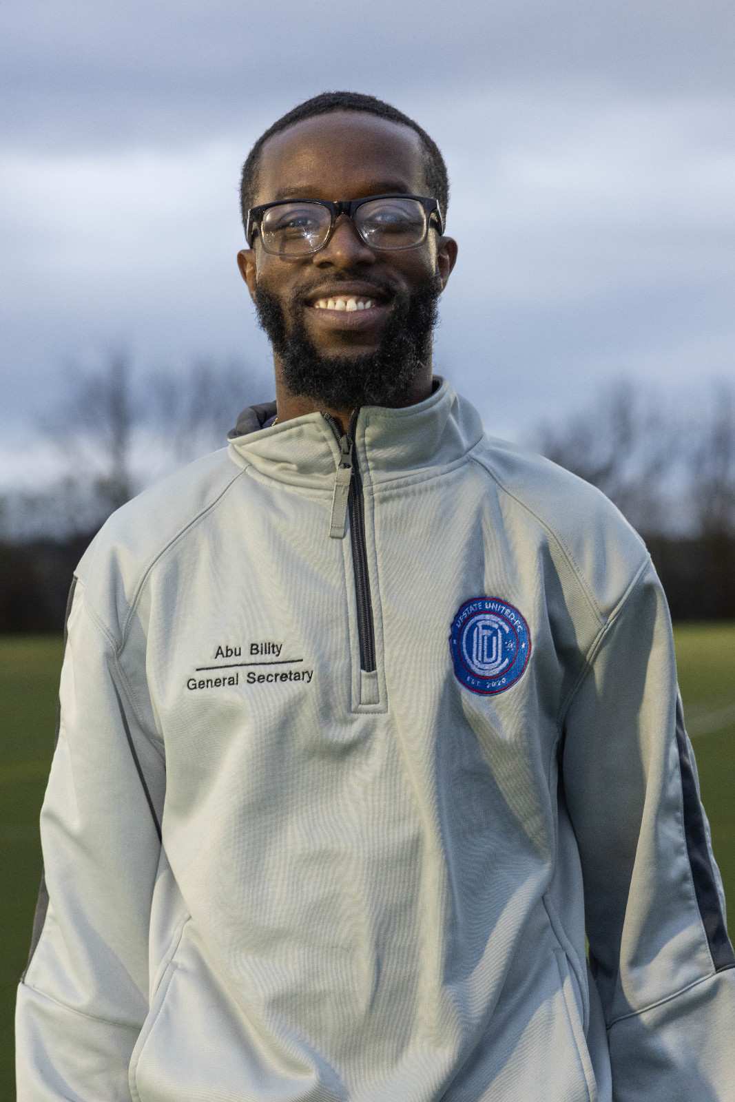 Smiling man with glasses and beard wearing a light gray zip-up jacket labeled 'Abu Billity General Secretary' and a round patch on the chest, standing outdoors.