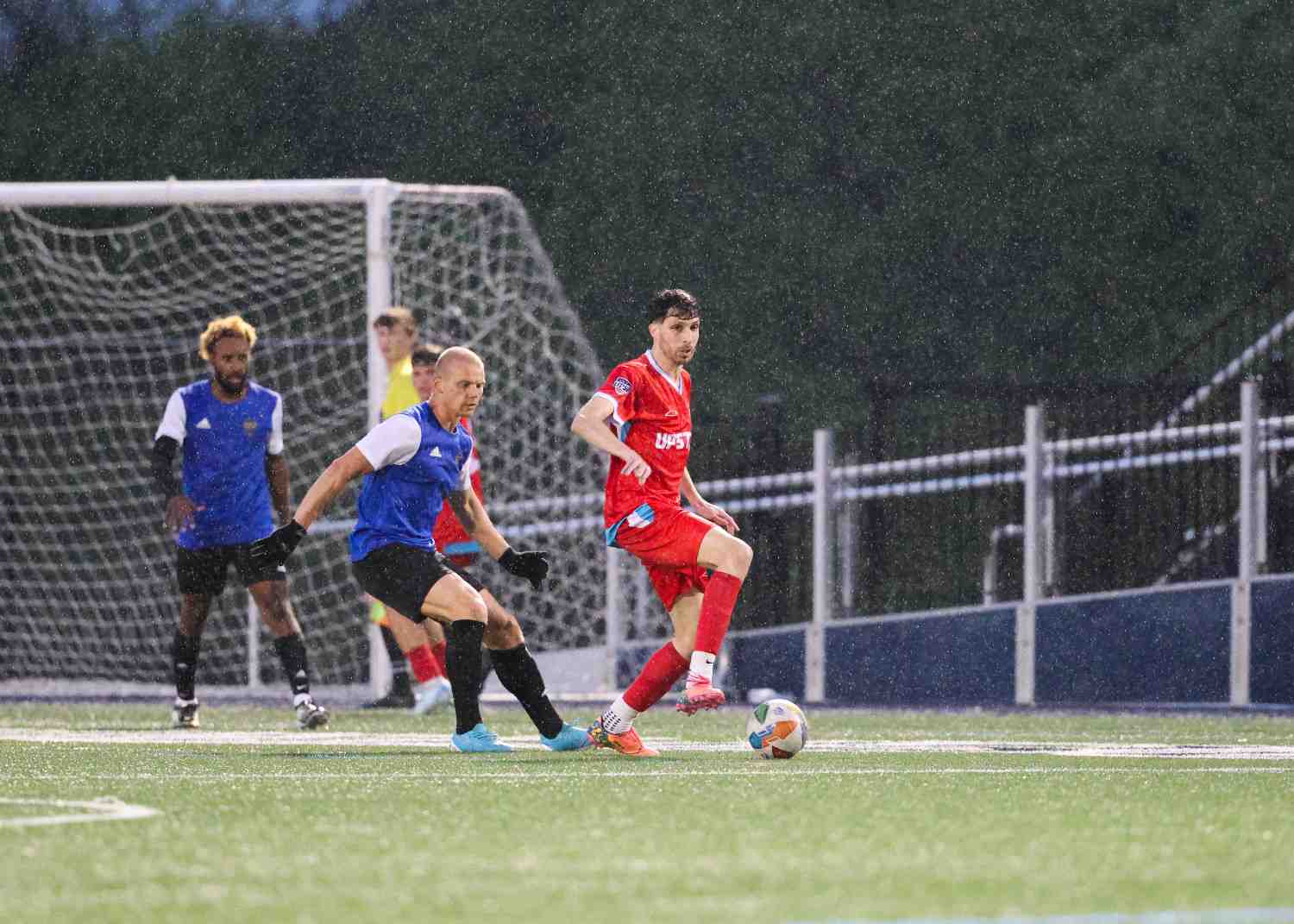Soccer players in action on a rainy field, with one player in red controlling the ball and players in blue defending near the goal.