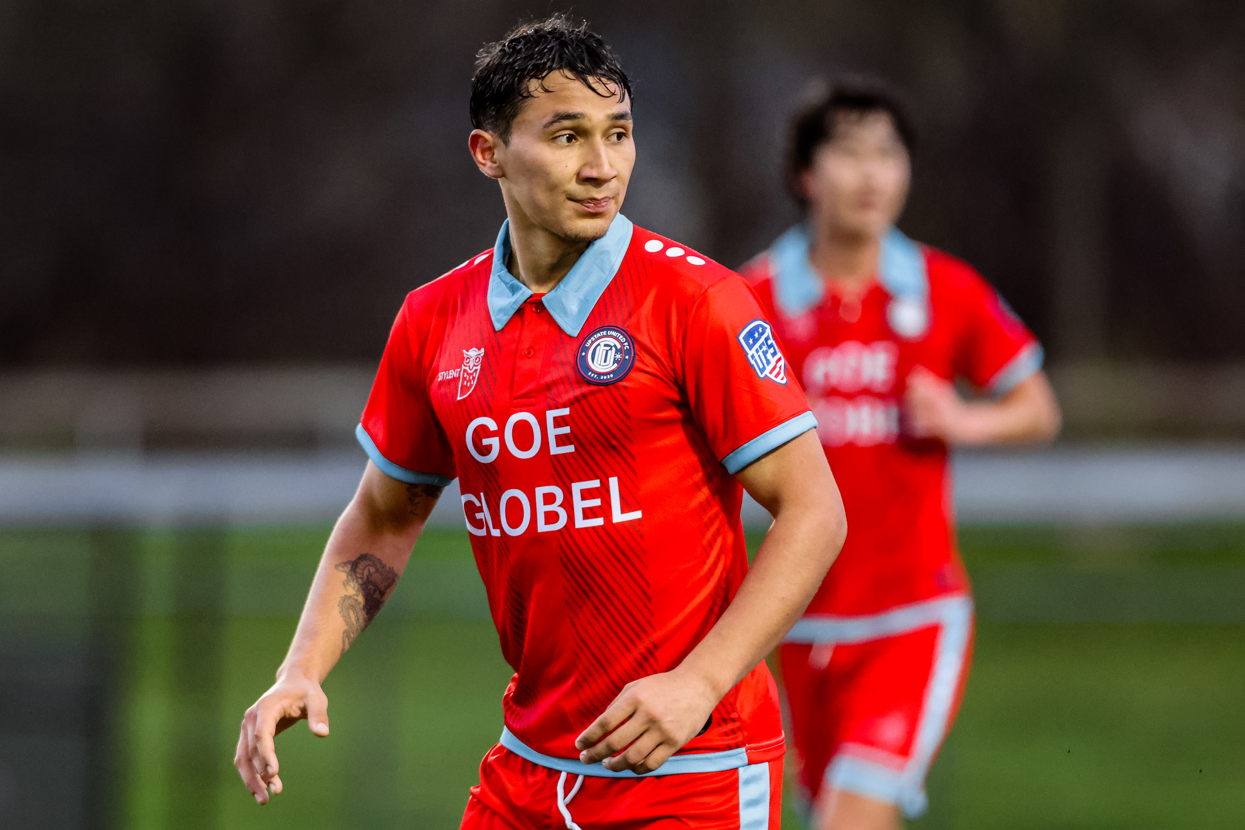 Male soccer player in red and blue uniform with 'GOE GLOBEL' on the jersey looking to the side on the field.