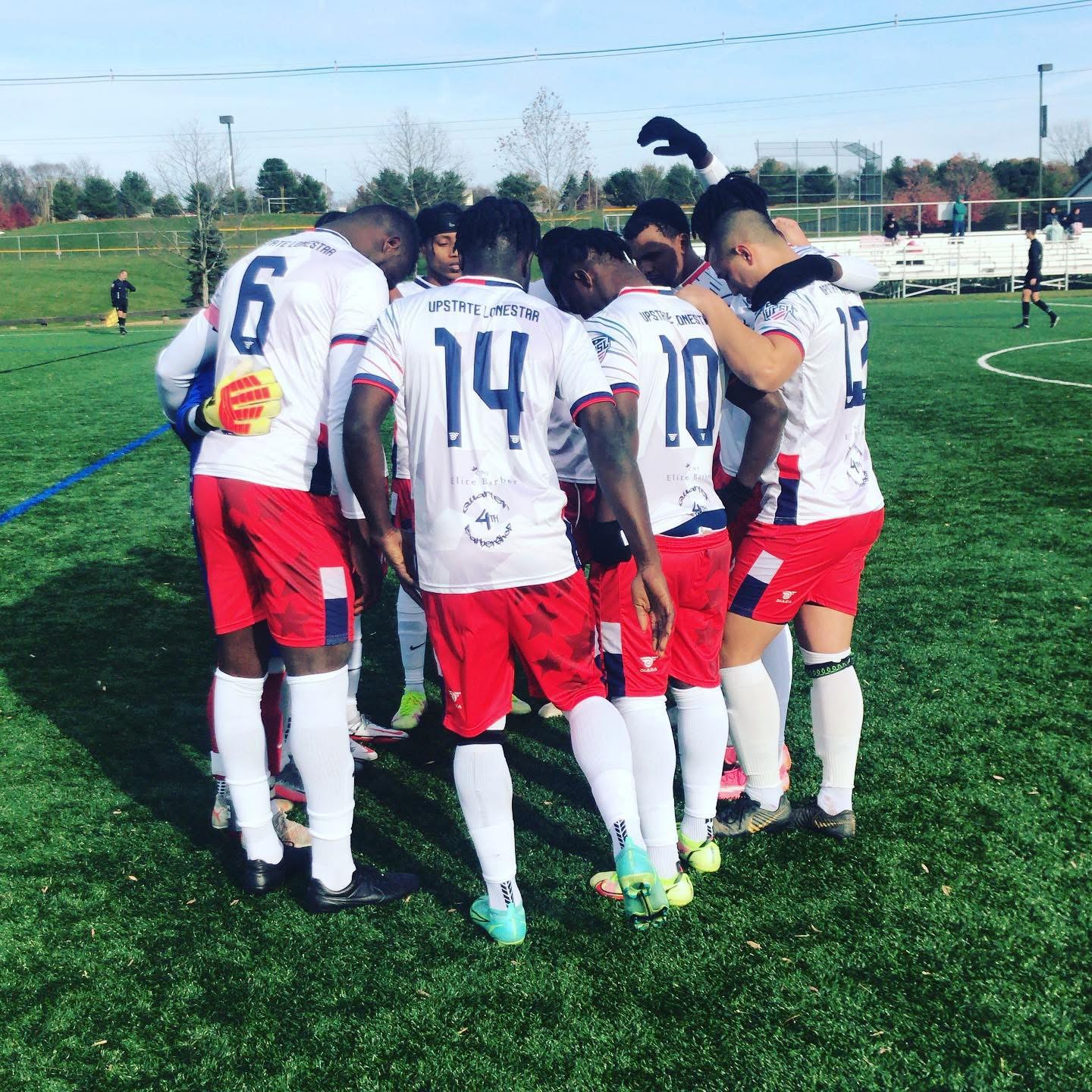 Soccer team in white jerseys and red shorts huddled together on a green field before a game.