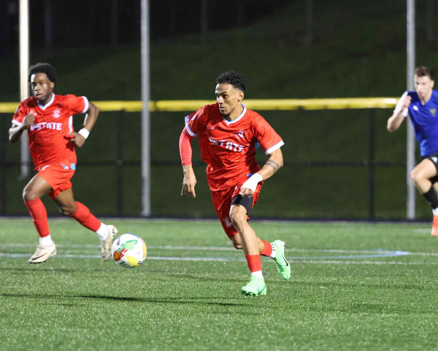 Soccer players in red uniforms running on a field at night toward a ball, with a player in blue chasing from behind.