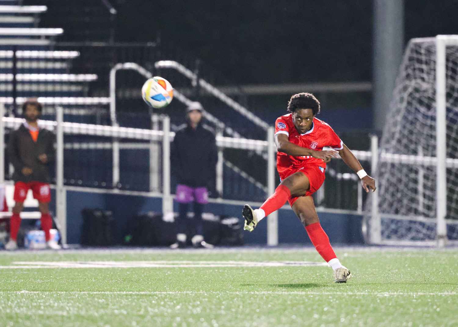 Soccer player in red uniform kicking a ball on a field with goal and spectators in the background.