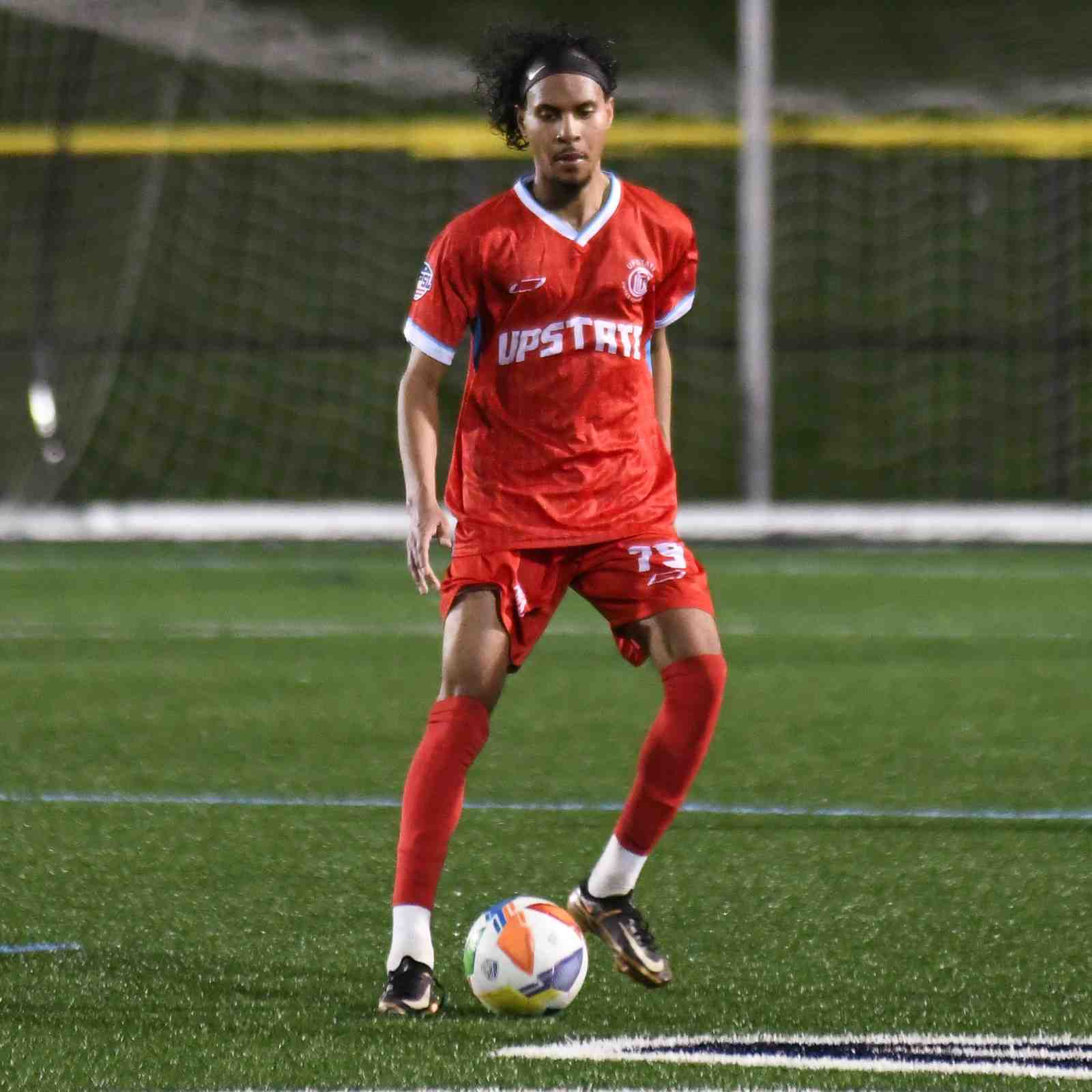 Soccer player in red Upstate United uniform controlling a soccer ball on a green field at night.