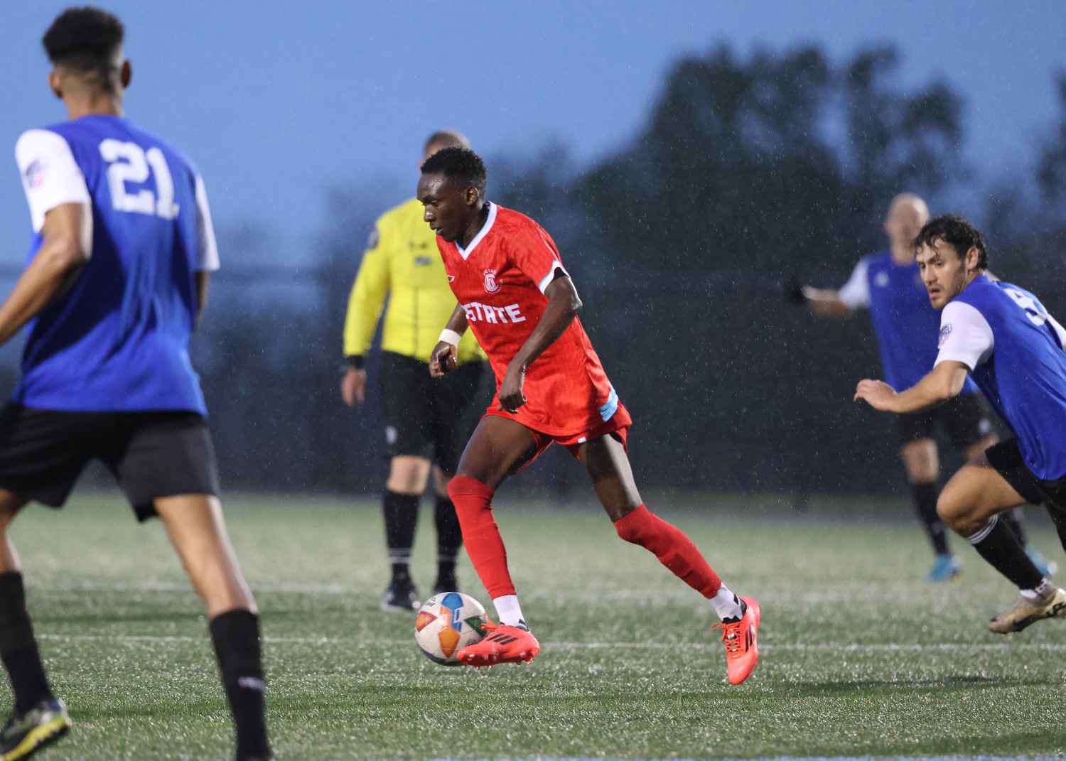 Soccer player in a red uniform dribbling a ball on a rainy field while opponents in blue jerseys approach.