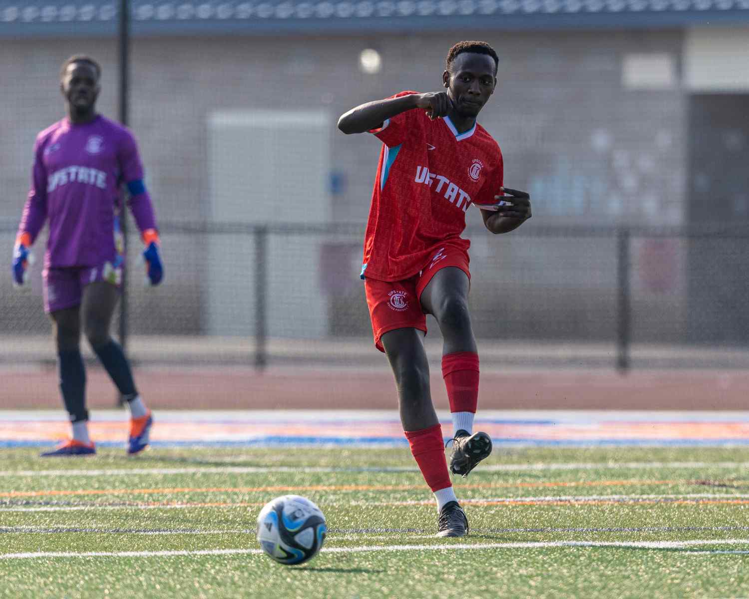 Soccer player in red uniform kicking a ball on a field with a goalkeeper in purple in the background.