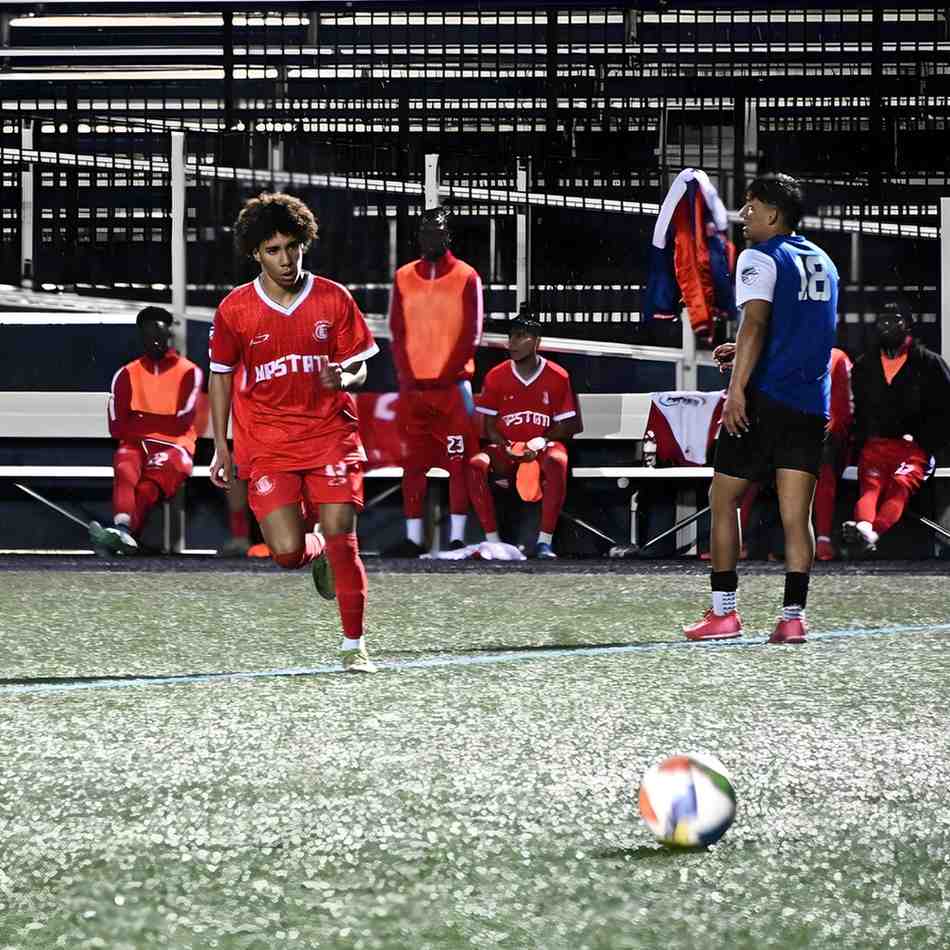 Soccer player in red uniform running on the field with a soccer ball nearby and teammates sitting on the bench in the background under stadium lights.