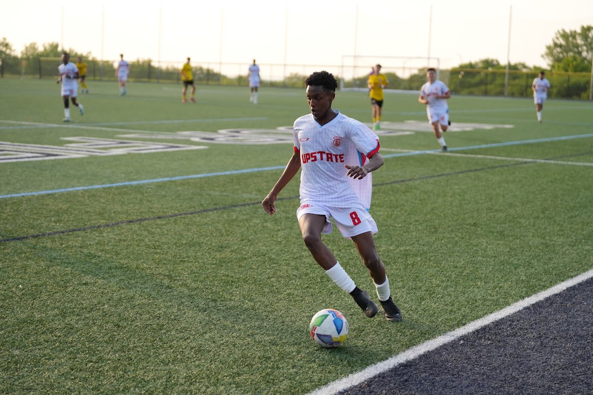 Soccer player wearing white Upstate jersey with number 8 controls a colorful soccer ball near the field sideline during a match.