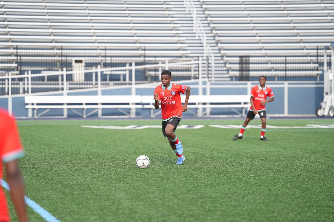Two boys in red soccer jerseys playing on a green field with empty bleachers in the background.