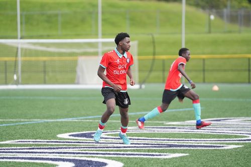 Two soccer players in red jerseys and black shorts running on a green field near a goalpost.