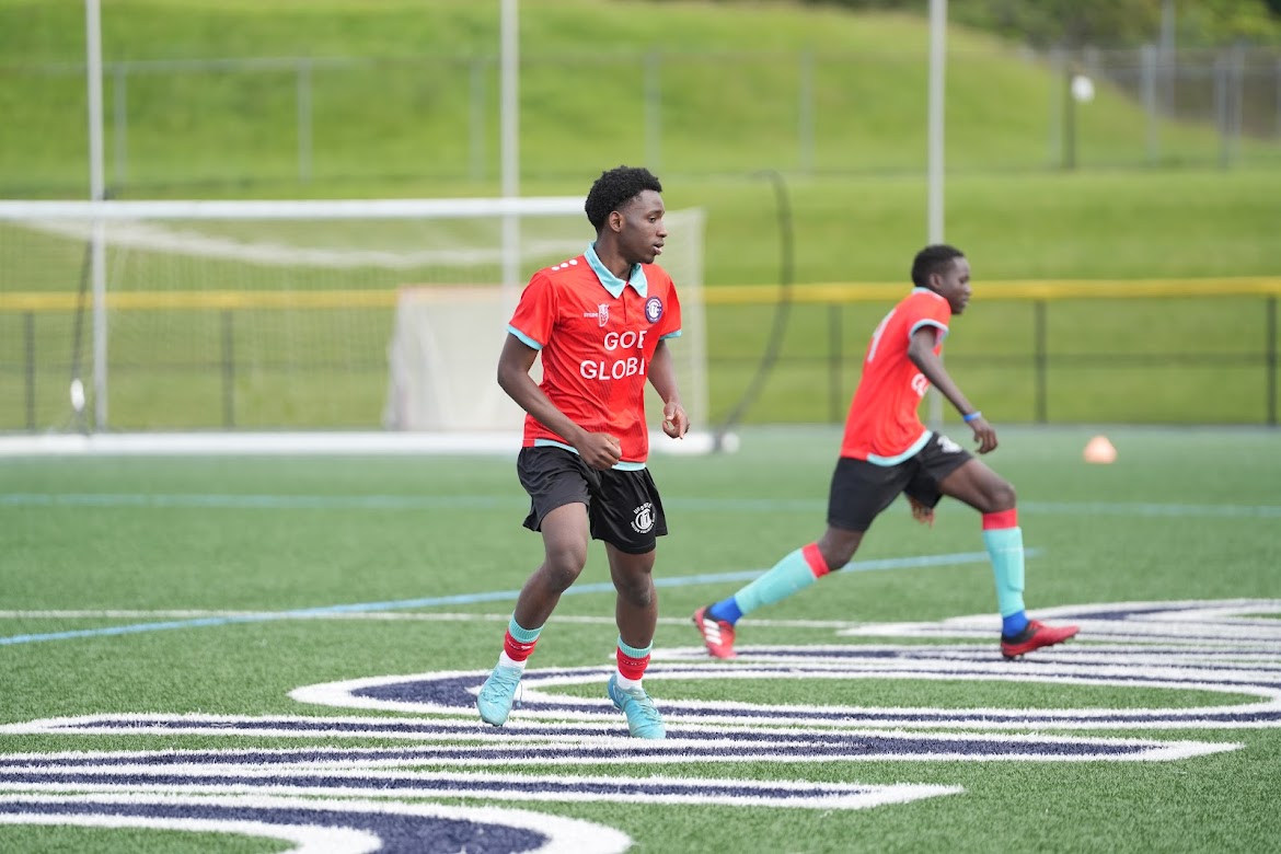 Two soccer players in red jerseys and black shorts running on a green field near a goalpost.