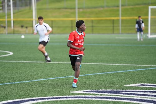 A soccer player in a red and light blue jersey running on a green field, with two other players and a goalpost in the background.