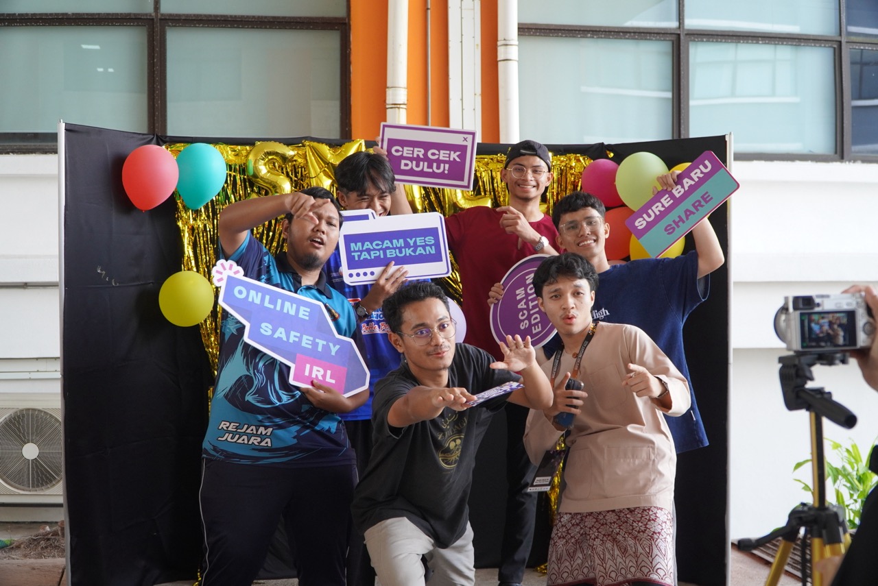 Six young men pose with colorful signs about online safety and scams at a photo booth with balloons.