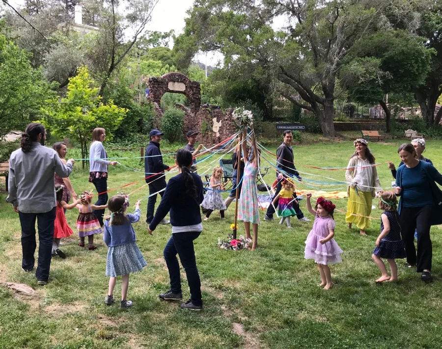 Parents and children gathered outdoors, dancing around a maypole with colorful ribbons during a community celebration.