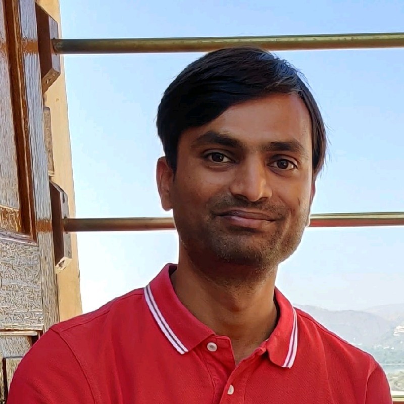 Man with short black hair wearing a red polo shirt smiling slightly with a wooden door and metal railing in the background against a clear blue sky.