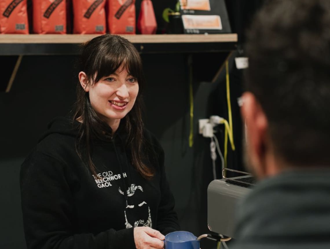 A staff member wearing a black hoodie jumper smiling and steaming milk at a coffee machine