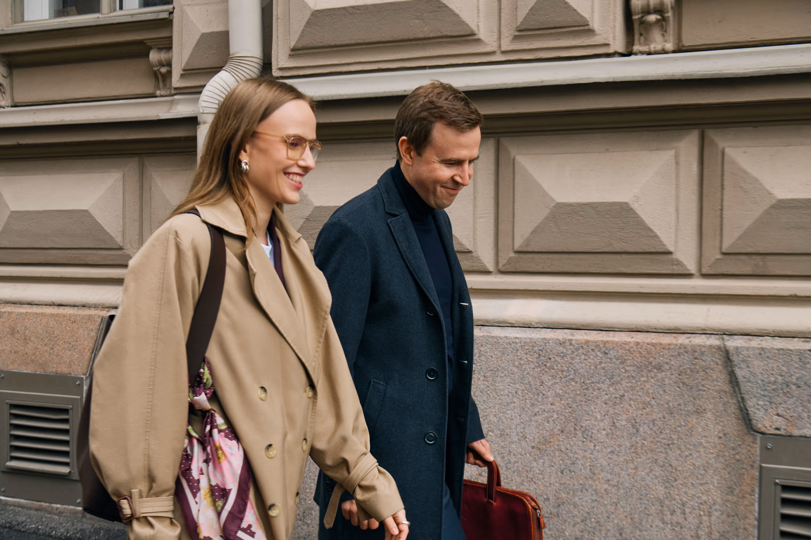 A smiling man in a dark coat carrying a briefcase and a woman in a beige trench coat walking together on a city sidewalk.