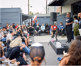 Outdoor event with speaker addressing seated audience near building and vehicle.