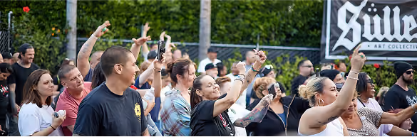 Crowd of people at an outdoor event raising their hands with a Sullen Art Collective banner in background.