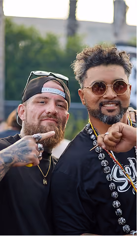 Two men posing outdoors; one points at the other who wears round sunglasses and a beaded necklace.