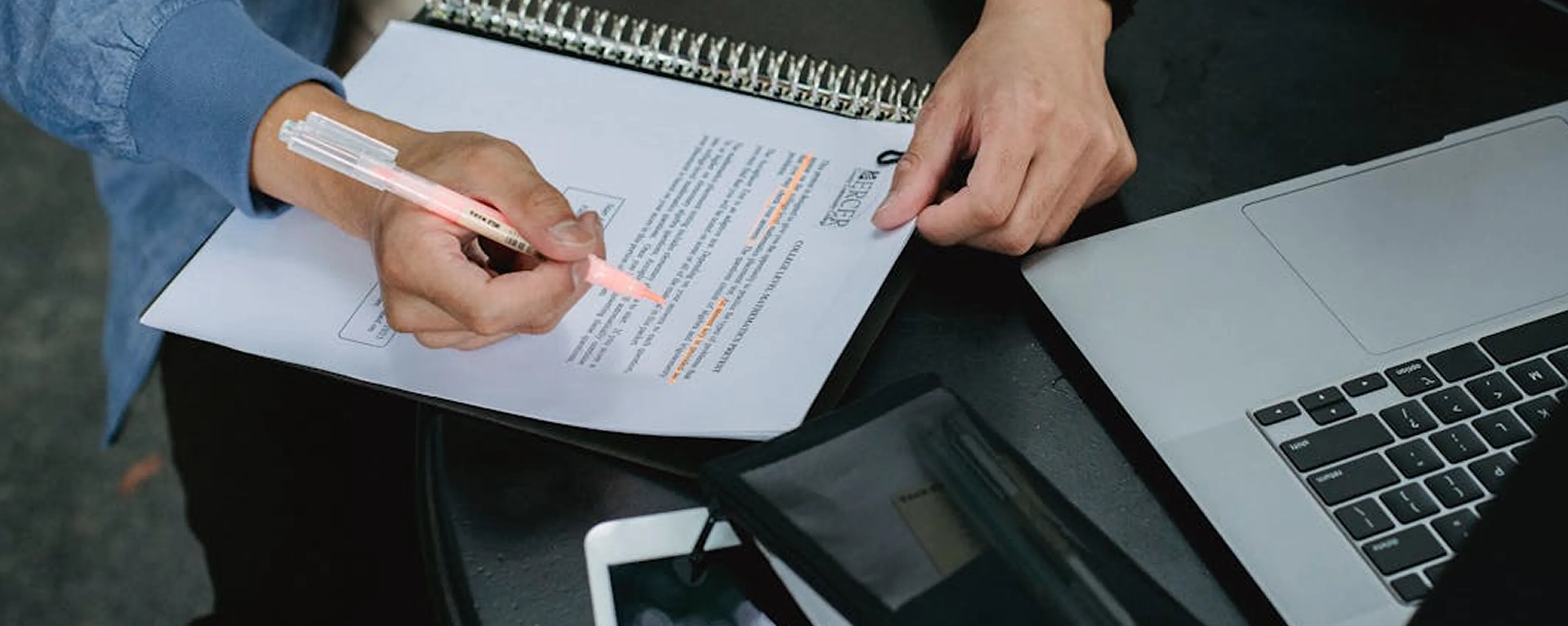 Person highlighting text on a paper document with a pink highlighter next to a laptop and a tablet on a black table.