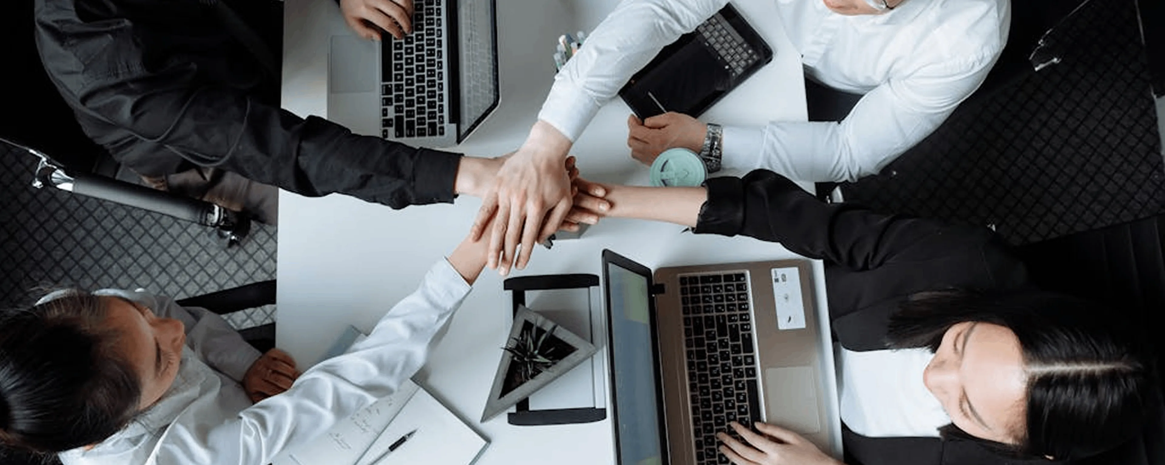 Four businesspeople seated around a table stacking hands in a teamwork gesture with laptops and notebooks on the table.