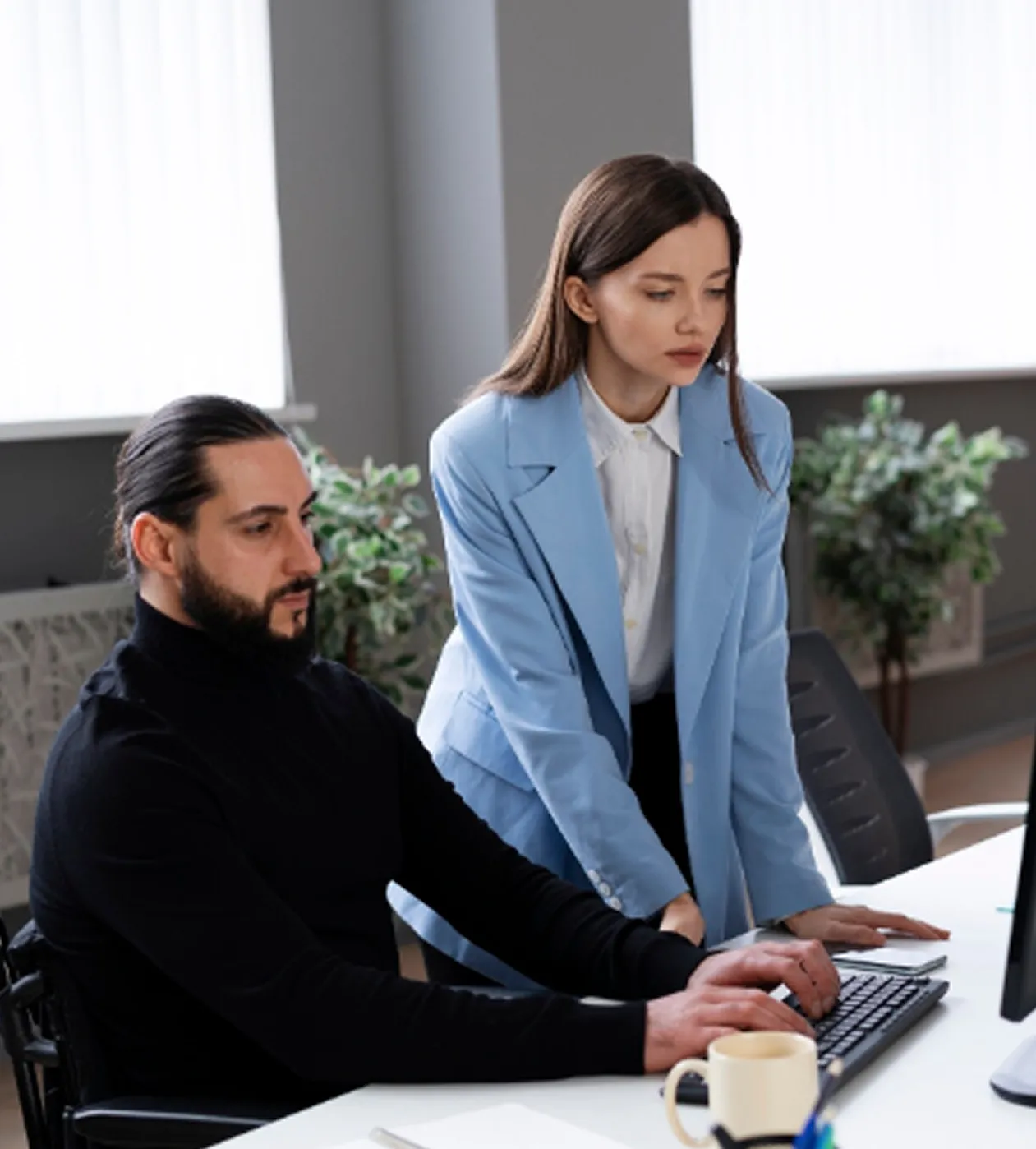 A man with a beard in a black turtleneck sitting and typing on a computer keyboard while a woman in a blue blazer leans over the desk watching.