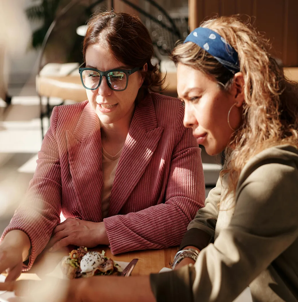 Two women sitting and looking at a smartphone on a wooden table with a plate of food.