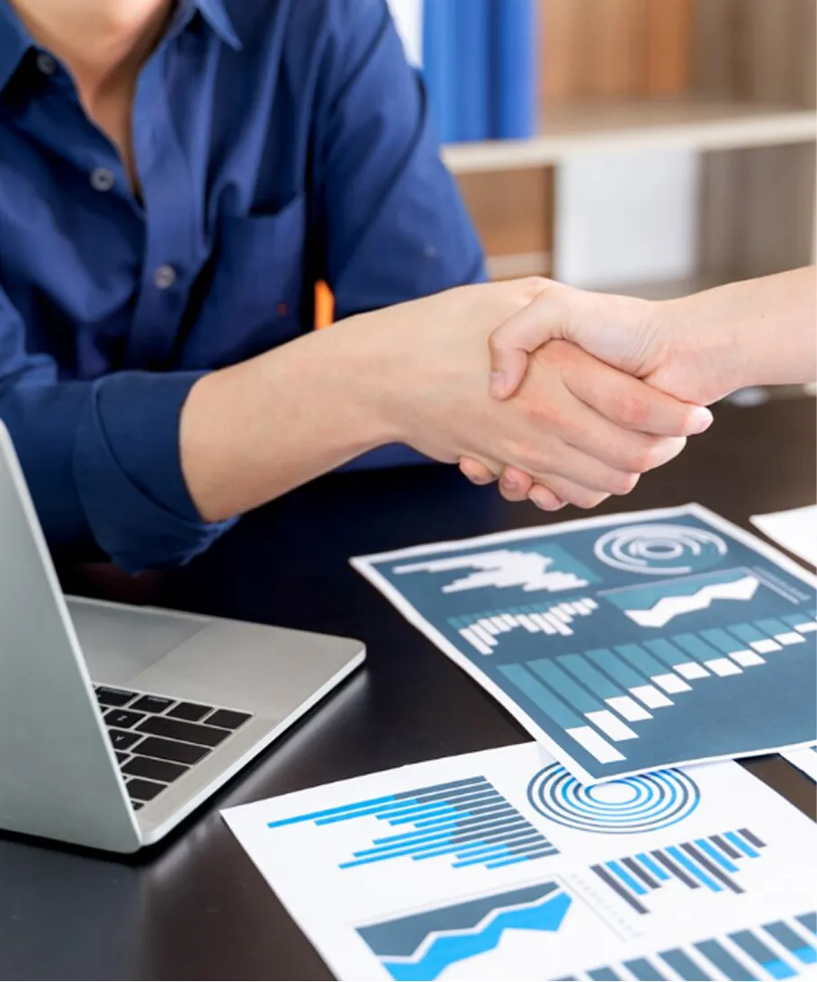 Two people shaking hands over a desk with a laptop and printed charts displaying data and graphs.