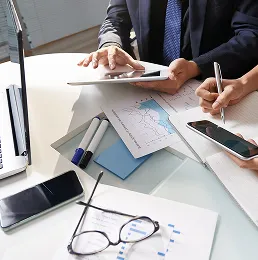 Two professionals reviewing financial charts and documents at a desk with smartphones, laptop, and glasses.