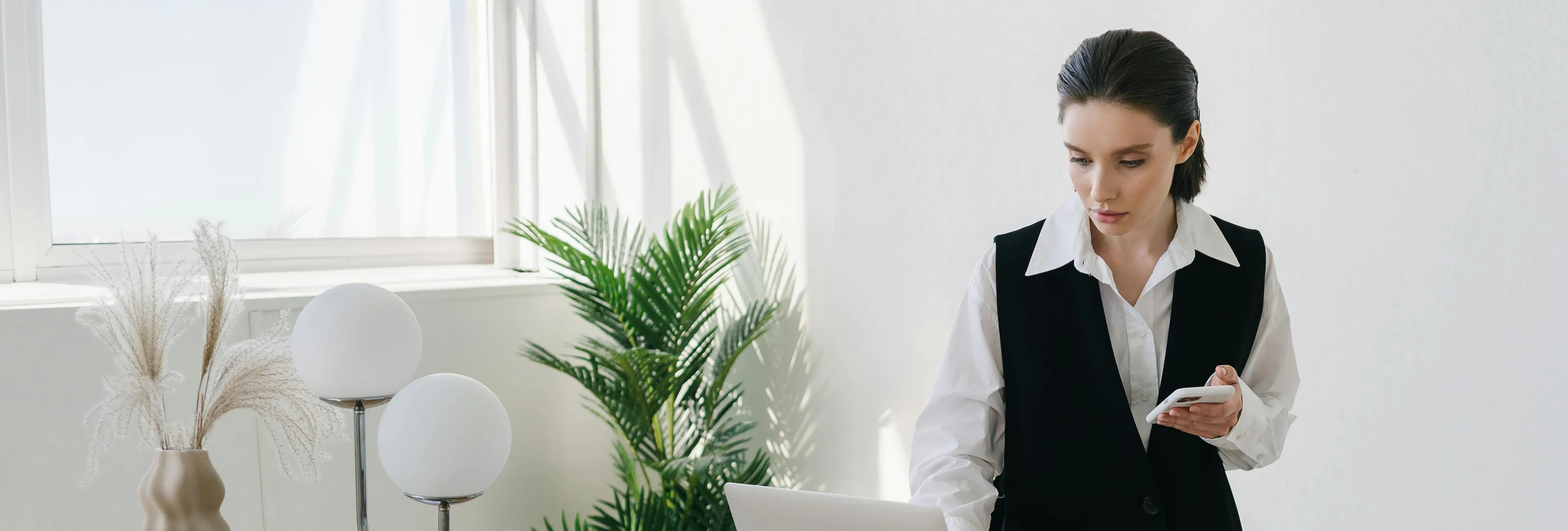 Woman in a white shirt and black vest looking at a laptop with a smartphone in hand in a bright room with plants and modern decor.
