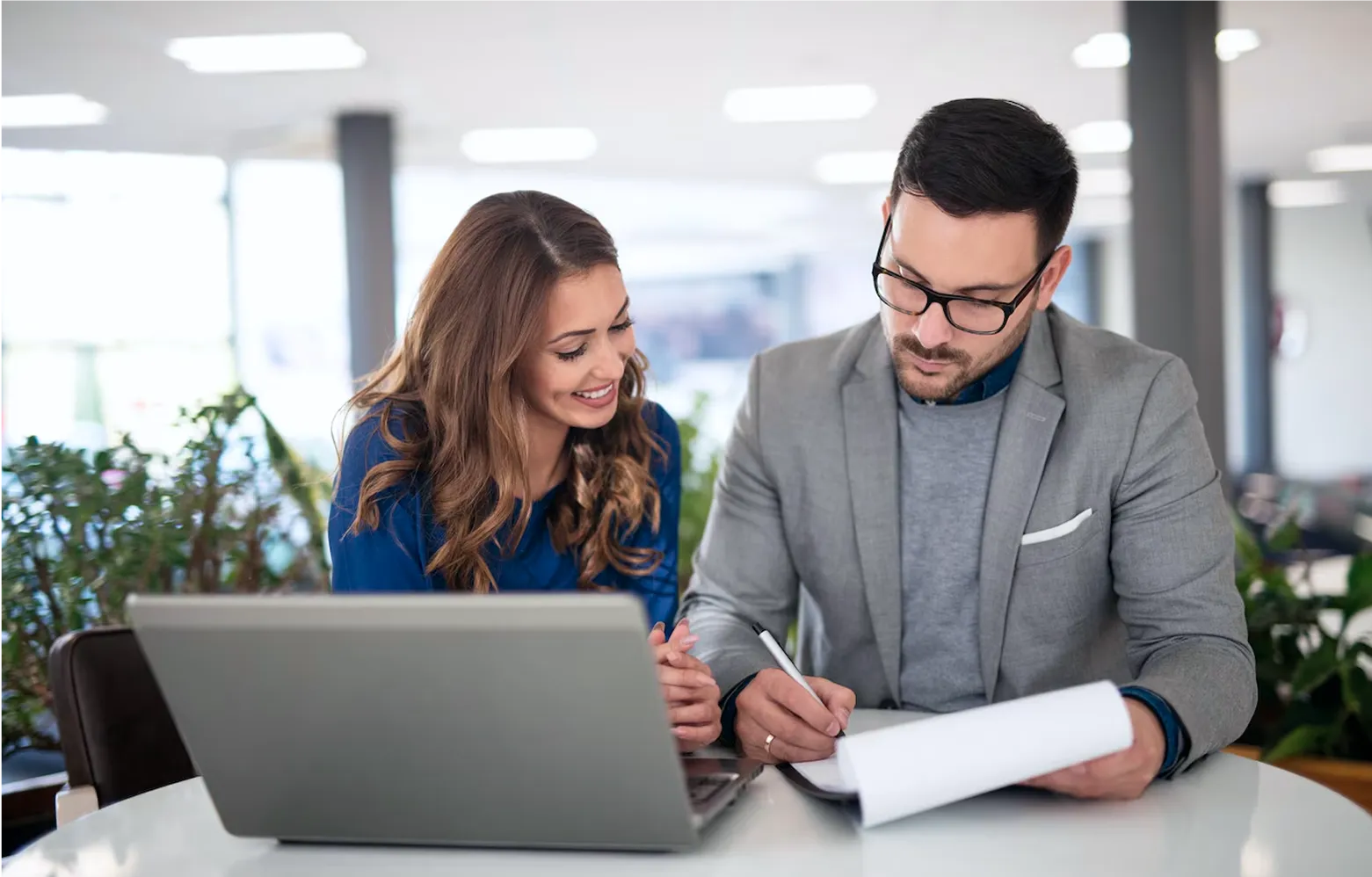 Man in gray suit and glasses writing on a clipboard while a woman in blue smiles beside him, with a laptop on the table.