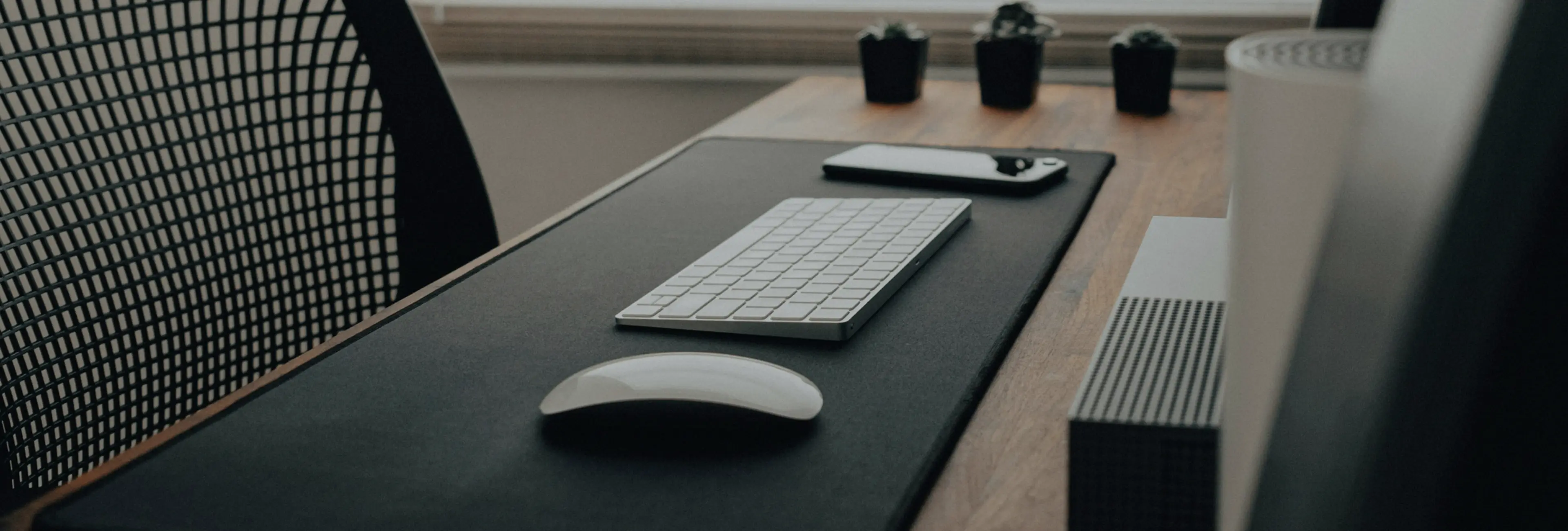 Minimalist workspace with a wireless keyboard, mouse, and smartphone on a desk with a mesh chair and small potted plants in the background.