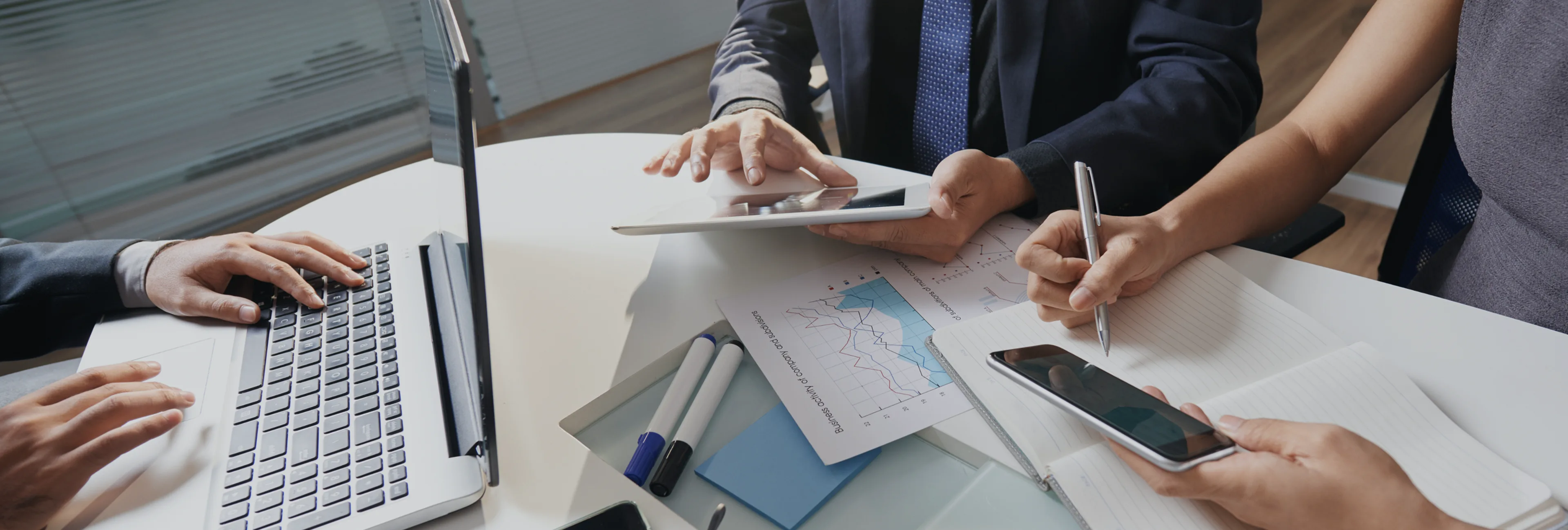 Three people at a round table working with a laptop, tablet, and smartphone alongside charts and notes.
