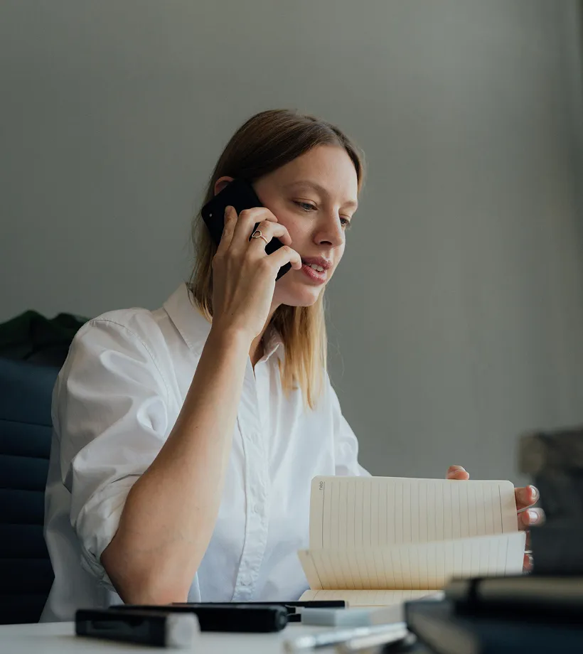 Woman in a white shirt talking on a phone while flipping through a notebook at a desk.
