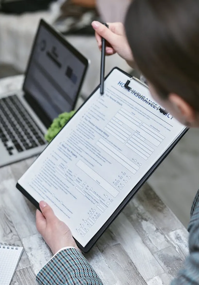 Person holding a clipboard with a home insurance policy form and pointing at it with a pen, laptop and notebook on table.