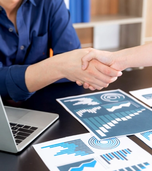 Two people shaking hands over a desk with a laptop and printed charts displaying graphs and data analysis.